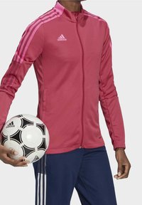 Chaqueta atlética roja con detalles en rosa y tres rayas en los hombros, combinada con pantalones azules. El modelo sostiene un balón de fútbol en blanco y negro.