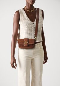 Borsa a tracolla in suede e pelle marrone con hardware dorato, forma rettangolare e tracolla regolabile. Indossata con un top senza maniche testurizzato di colore crema.