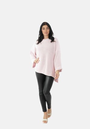 Kvinde med langt mørkt hår iført en lys pink oversized striktrøje, sorte læderbukser og hvide åbne tå-sandaler, stående og poserende.