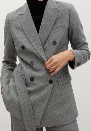 Personne portant un blazer gris croisé avec des boutons noirs sur un haut noir, ajustant le blazer d'une main dans la poche avant.
