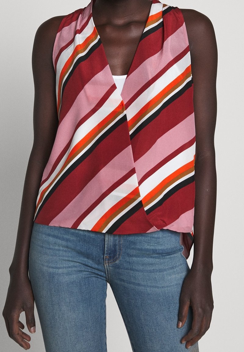 Blouse sans manches style portefeuille avec des rayures diagonales rouges, roses, oranges, blanches, noires et brunes portée avec un jean bleu.