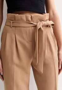 Pantalon taille haute beige avec une large ceinture nouée. Présente des plis à l'avant et des poches latérales. Tissu lisse et texturé.