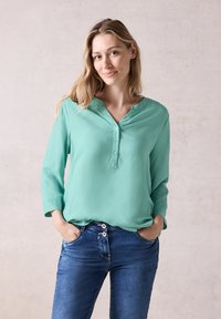 Helles türkisfarbenes Langarmbluse mit V-Ausschnitt und Knopfdetails. Kombiniert mit dunkelblauen Jeans aus Denim, die einen stilvollen Knopfverschluss auf der Vorderseite haben.