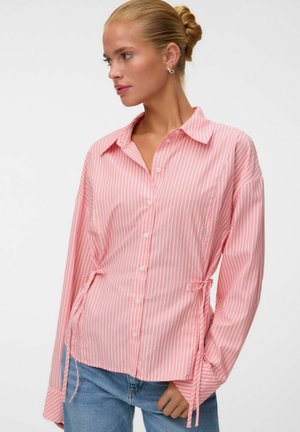 Femme aux cheveux blonds attachés en chignon, portant une chemise rayée rose et blanche à boutons avec des liens à la taille et un jean bleu sur un fond uni.