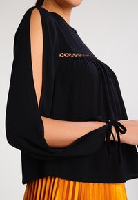 Blusa nera con maniche lunghe, caratterizzata da aperture sulle spalle e dettagli con legature ai polsi. Tessuto leggero con una vestibilità ampia.