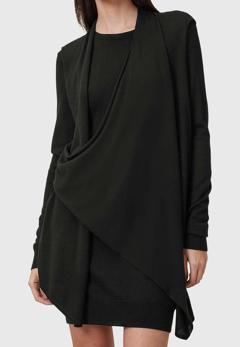 Femme portant une robe en maille noire à manches longues avec un col châle noir drapé par-dessus.