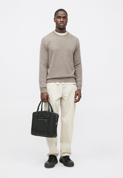 Gaiši brūns vilnas džemperis pār baltu kreklu, krēmkrāsas bikses un melni sporta apavi. Viņš tur melnu strukturētu somu ar augšējiem rokturiem un rāvējslēdzēja detaļu.