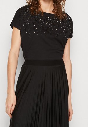 Vrouw die een zwart shirt met korte mouwen draagt, versierd met gouden studs op de borst, gecombineerd met een zwart geplooide rok met hoge taille.