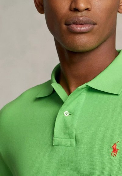 Primo piano di un giovane uomo che indossa una polo verde brillante con un piccolo logo rosso, dalla pelle liscia e un'espressione neutra.