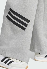 Szare dresowe spodnie mają trzy czarne paski po lewej stronie. Model nosi białe sneakersy z czarnymi i szarymi akcentami.