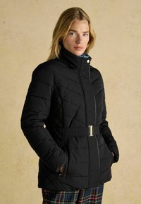 Melnā izšūta pūkaina jaka ar augstu apkakli, jostasvietu un sānu kabatām. Materiāls izskatās gluds ar matētu apdari.