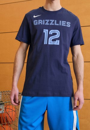 Persoon die een marineblauw shirt met 'Grizzlies' en nummer 12 draagt, samen met blauwe basketbalshorts, staat naast een winkelwagen vol met basketbalballen.