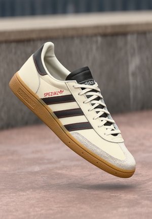 Valkoinen Adidas Spezial -tennari mustilla raidoilla, kumipohjalla ja punaisella logoyksityiskohdalla, kuvattuna ilmassa sumeaa kaupunkitaustaa vasten.