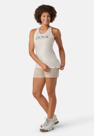 Top sans manches beige clair avec broderies florales, associé à un short beige et des chaussures de randonnée blanches avec des accents verts.