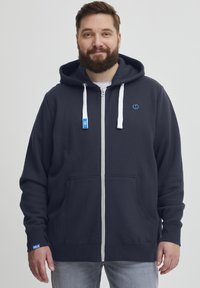Mornarica zip-up dukserica izrađena od mekog materijala, s bijelim vezicama, kenguru džepovima i malim logotipom na prsima. Osvježeni rukavi u svijetloplavoj boji.