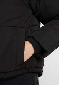 Černá péřová bunda s prošívanou strukturou, která má přední kapsu a žebrované manžety. Lehký materiál s hladkým povrchem.