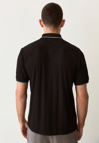 Zwart poloshirt met korte mouwen en een gestructureerd diamantenpatroon, contrasterende grijze kraag en manchetkappen. Achteraanzicht toont een gebogen zoom.