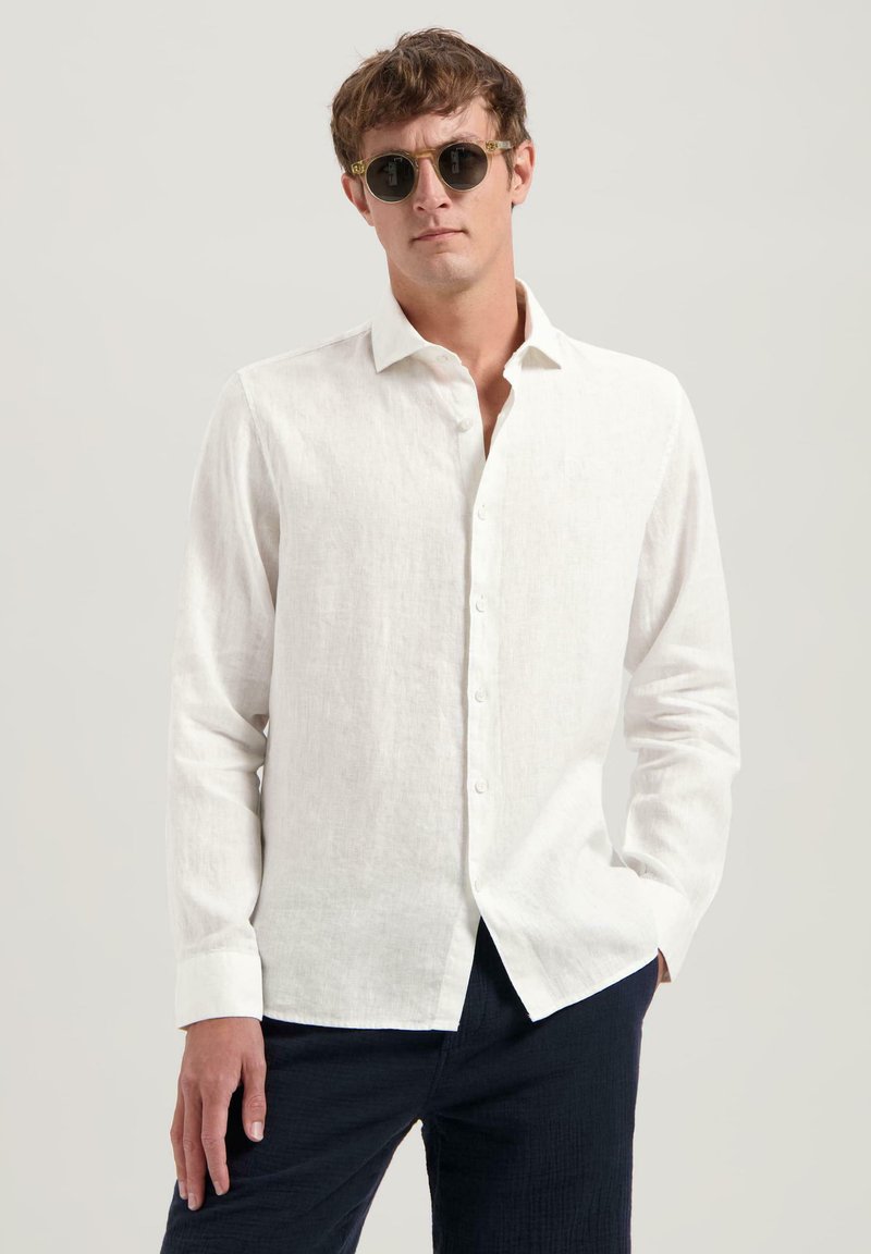 Camicia bianca in lino a maniche lunghe con bottoni davanti e dettagli ai polsini, abbinata a pantaloni scuri; modella che indossa occhiali da sole tondi.