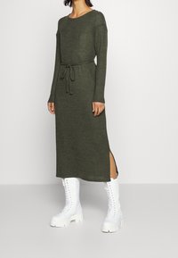 Lång, olivgrön stickad klänning med långa ärmar, dragsko i midjan och slitsar på sidorna, ihop med vita snörade kängor.