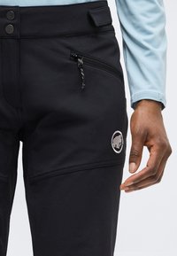 Prim-plan cu pantaloni negri cu buzunar cu fermoar și logo mamut pe coapsă, purtați cu o cămașă cu mâneci lungi bleu deschis și mâna dreaptă vizibilă.