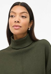 Pull ras du cou vert olive avec texture côtelée et ourlet fendu. Boucles d'oreilles pendantes en argent complètent le design. Style épuré et minimaliste.