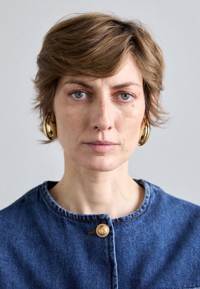 Traper jakna s ovalnim izrezom, s zatvaranjem na zlatne gumbe. Model ima kratku, valovitu kosu i nosi velike zlatne naušnice.
