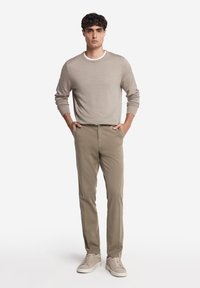 Maglione a maglia beige chiaro con scollo rotondo, abbinato a chinos beige chiaro e sneakers beige. Il modello è in piedi con le mani nelle tasche.