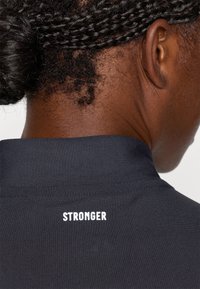 Svart atletisk topp med høy krage og ordet "STRONGER" i hvit skrift bak. Glatt stoff med en tettsittende design.
