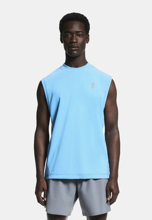 Hombre con camiseta deportiva sin mangas azul claro y pantalones cortos grises, de pie mirando hacia adelante sobre un fondo liso.