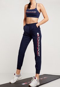 Navy sportieve crop top en joggings met een wit en rood "TOMMYSPORT" logo. Model draagt witte sneakers met accenten. Soepele stoftextuur.