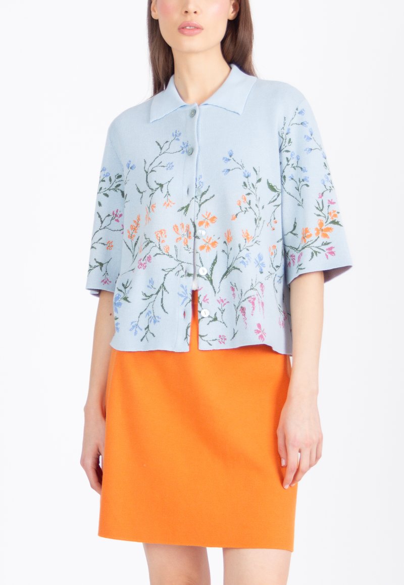 Žena v svetlomodrom zapínacom kardigane s viacfarebnou kvetinovou výšivkou a jasne oranžovej sukni, stojaca na bielom pozadí.