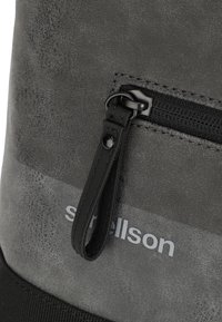 Material de mochila texturizada gris con un cierre negro liso, detalle de cuero y un logo plateado en la parte frontal, enfatizando la durabilidad y el diseño.