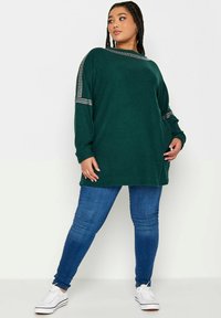 Maglione verde oversized in tessuto texturizzato, con scollatura a barca e spalle decorate, abbinato a jeans skinny blu e sneakers bianche.