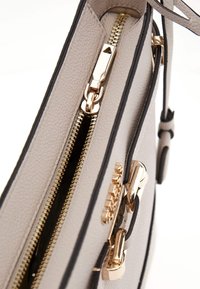 Borsa a mano in pelle grigia chiara con dettagli neri, hardware dorato, doppie zip e superficie texturizzata. Gli accenti includono zip pulls angolari.