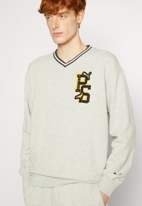 Grauer Pullover mit V-Ausschnitt und kontrastierendem dunklen Halsbereich, ausgestattet mit einem gelb-schwarzen Logo, das die Buchstaben "PS" und einen grafischen Akzent zeigt.