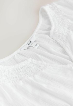 Baltas viršus, pagamintas iš lengvos, tekstūruotos medžiagos, su susmulkintais detalėmis aplink kaklo liniją. Matomi etiketės su prekės ženklu ir priežiūros informacija.