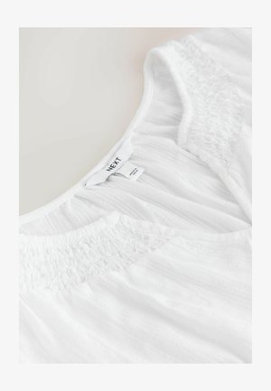 Top bianco realizzato in tessuto leggero e strutturato con dettagli arricciati intorno al collo. Etichette visibili con informazioni sul marchio e sulla cura.