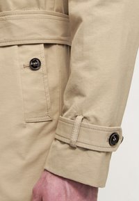 Manica di trench beige con dettaglio di bottone e cinturino regolabile, realizzata in tessuto morbido con una finitura pulita e sartoriale.