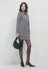 Abito grigio a maniche lunghe con colletto, abbinato a collant neri trasparenti e tacchi a punta neri. Borsa nera con un design curvo.