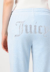 Gaiši zili sporta bikses, kas izgatavotas no mīkstas materiāla, aizmugurē rotātas ar lielu, mirdzošu rhinestones dizainu ar vārdu "Juicy".