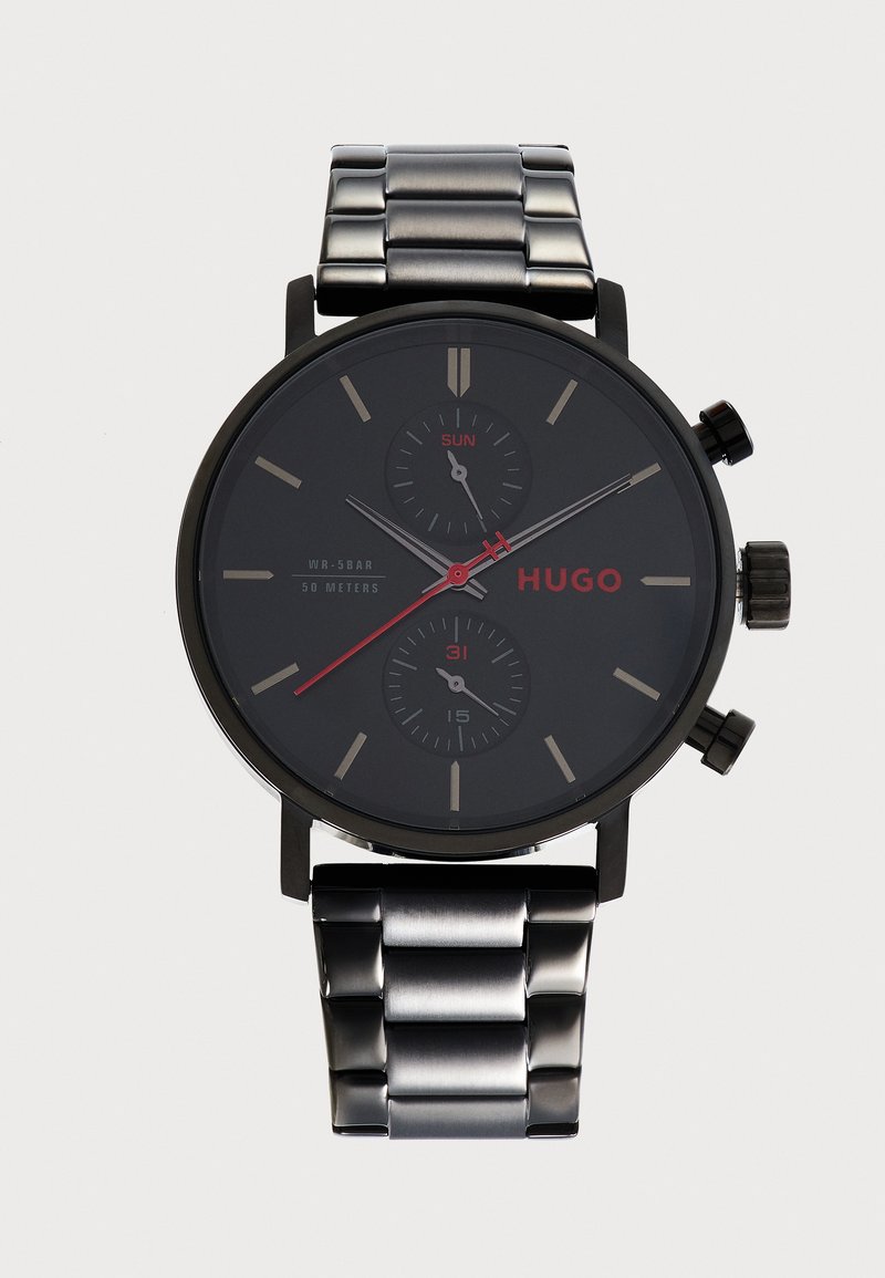 Montre-bracelet en métal noir avec des index heures argentés, une trotteuse rouge, deux sous-cadrans indiquant le jour et la date, et un logo HUGO rouge sur le cadran.