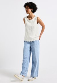 Vit ärmlös topp med hålbroderi, vågig fåll; matchad med ljusblå vida jeans och vita sneakers. Texturerad tyg.