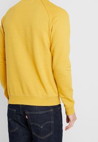 Gul sweatshirt med ribbad nederkant och mudd, med en slät textur. Bärs tillsammans med mörkblå jeans som har sömmar och fickor.