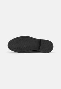 Semelle de chaussure noire avec un motif en zigzag texturé et des bords arrondis. Logo estampé et matériau confortable et flexible.