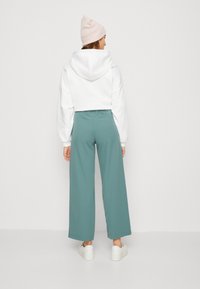 Biely skrátený mikin s kapucňou a balónovými rukávmi skombinovaný s teal širokými nohavicami. Model má na sebe biele tenisky a svetloružovú čiapku.