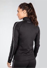Chaqueta atlética negra con cuello alto, rayas blancas a los lados, un diseño ajustado y una textura de tela suave. Logo visible en la parte posterior.
