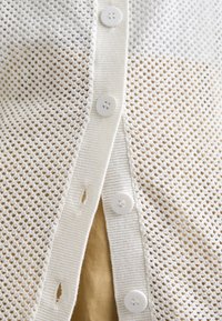 Cardigan bianco lavorato a maglia con un design a rete, caratterizzato da chiusure a bottone distanziate uniformemente e una texture liscia, sovrapposto a uno sfondo marrone.
