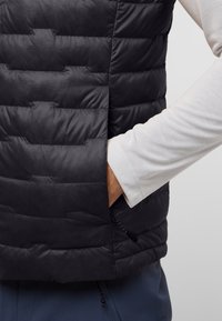 Schwarze isolierte Weste mit gestepptem Muster, ausgestattet mit einer Seitentasche und schlankem, leichtem Design. Getragen über einem hellen Langarmshirt.