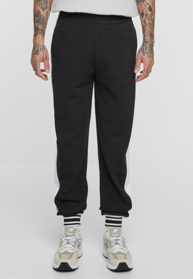 Schwarze Sweatpants mit einem weißen Seitenstreifen, aus weichem Material. Mit geripptem Knöchel-Design, kombiniert mit grauen und beigen Sneakers und gestreiften Socken.