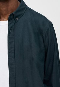 Donkergroen blouse met knopen, gemaakt van glad stof, voorzien van een puntige kraag en een enkele borstzak, gelegd over een wit t-shirt.